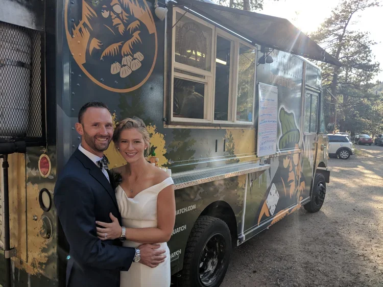 A couple dressed in formal attire standing in front of a food truck at sunset.