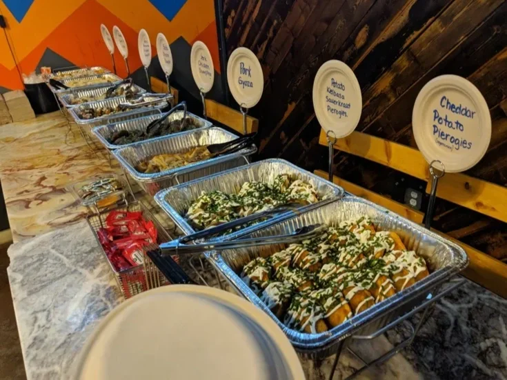 Buffet table with several trays of food, including nachos with cheese, and labels for different dishes like pork gyoza, potato kabobs, and cheddar potato pierogies.