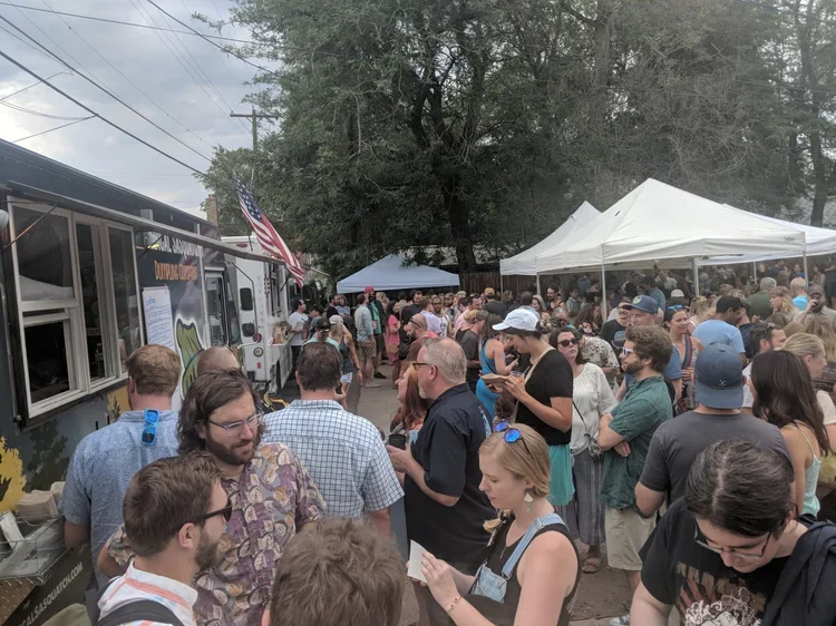Crowd of people gathering outdoors at a food truck event with white tents and trees in the background.
