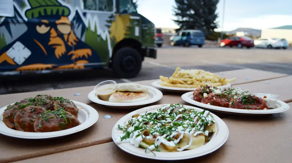 Four plates of food including meat with gravy, fried potato chips topped with cheese and herbs, a breakfast sandwich with eggs and bacon, and a plate of spaghetti with tomato sauce and herbs, outdoors on a table with a parking lot and a painted truck in the background.