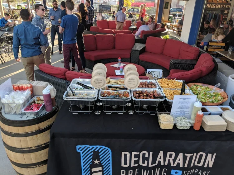 Buffet table with assorted food and drinks at Declaration Brewing company, in front of a seating area with red couches and people socializing outdoors.