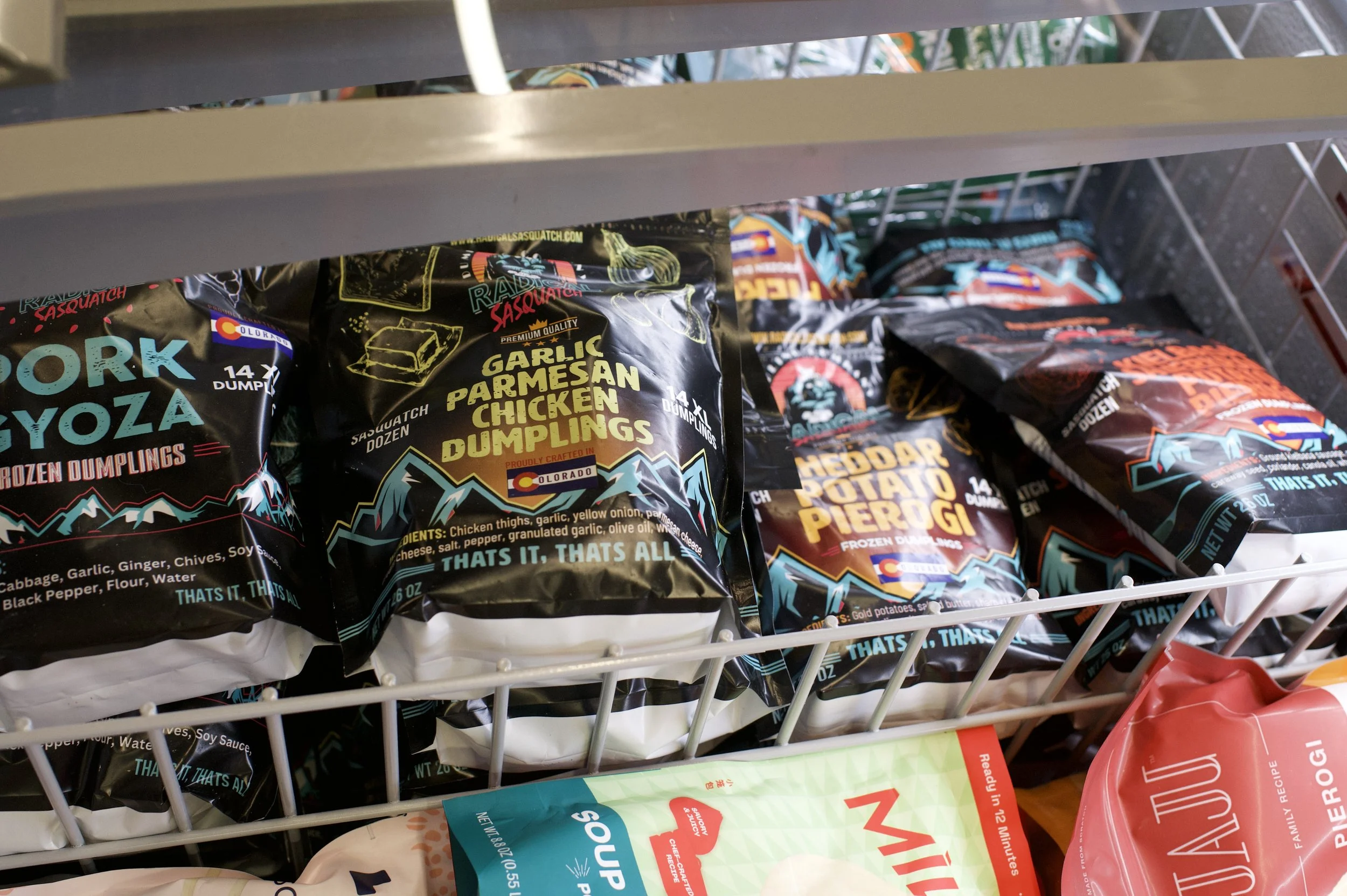 Frozen dumplings in various flavors on a grocery store shelf, including garlic parmesan chicken, cheddar potato pierogi, and pork gyoza.