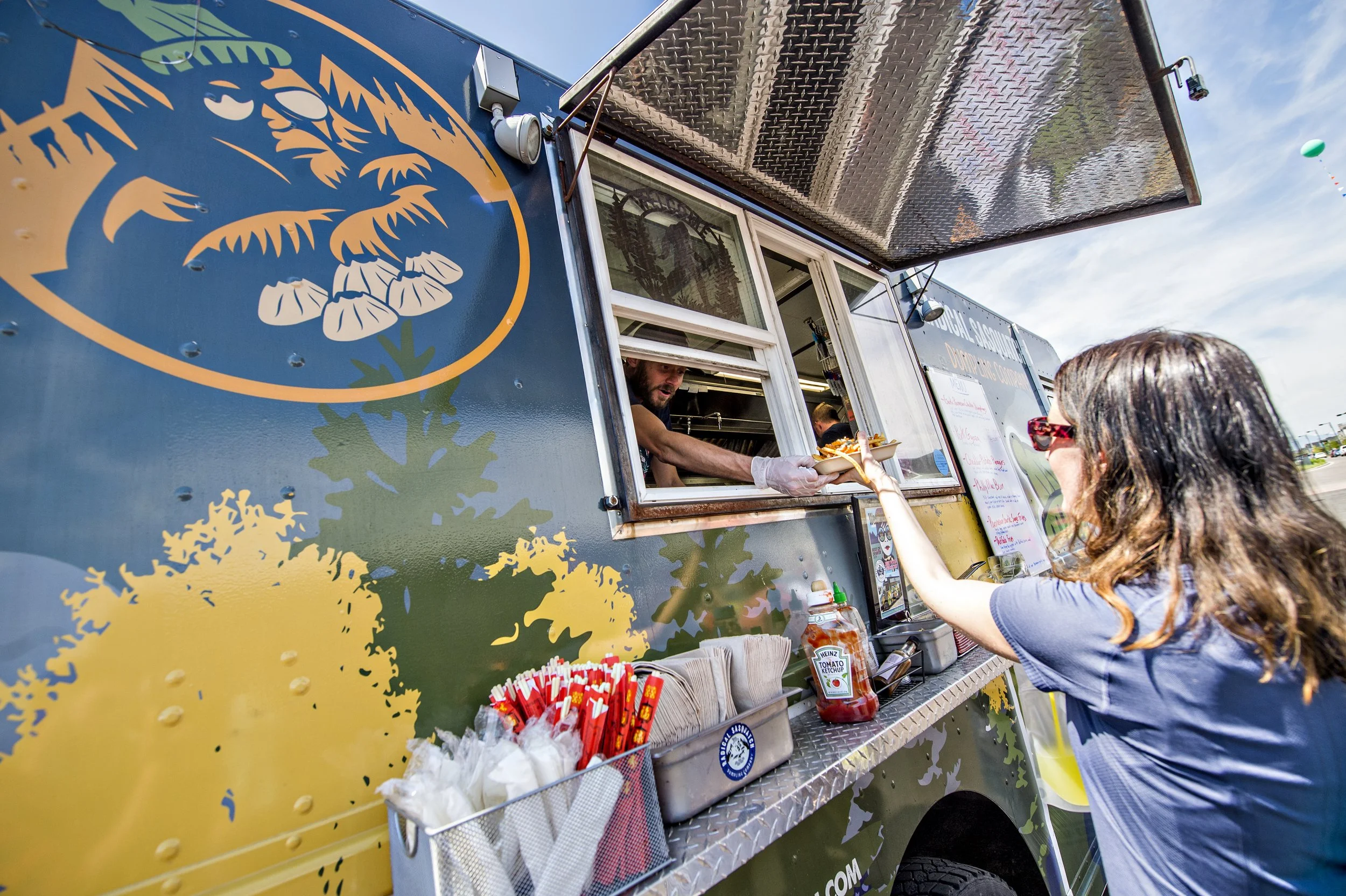 A woman is ordering food from a food truck that has a colorful foliage logo. The woman is wearing sunglasses and a gray shirt, and the food truck is serving a food item through a window.