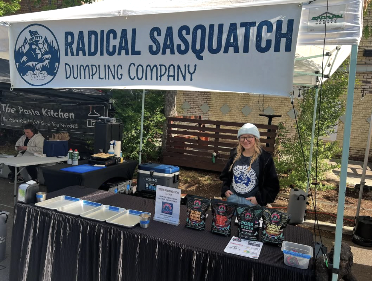 A person standing behind a table at a food booth named Radical Sasquatch Dumpling Company, with product packages displayed on the table, at an outdoor event.