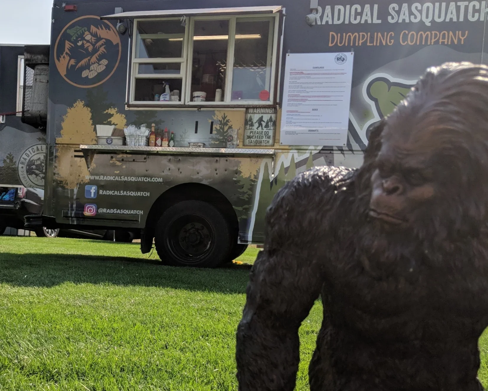 A food truck named 'Radical Sasquatch Dumpling Company' parked on a grassy area, with a Sasquatch figure in the foreground. The truck has social media handles on the side, a window open with some items visible inside, and a menu posted on the side.