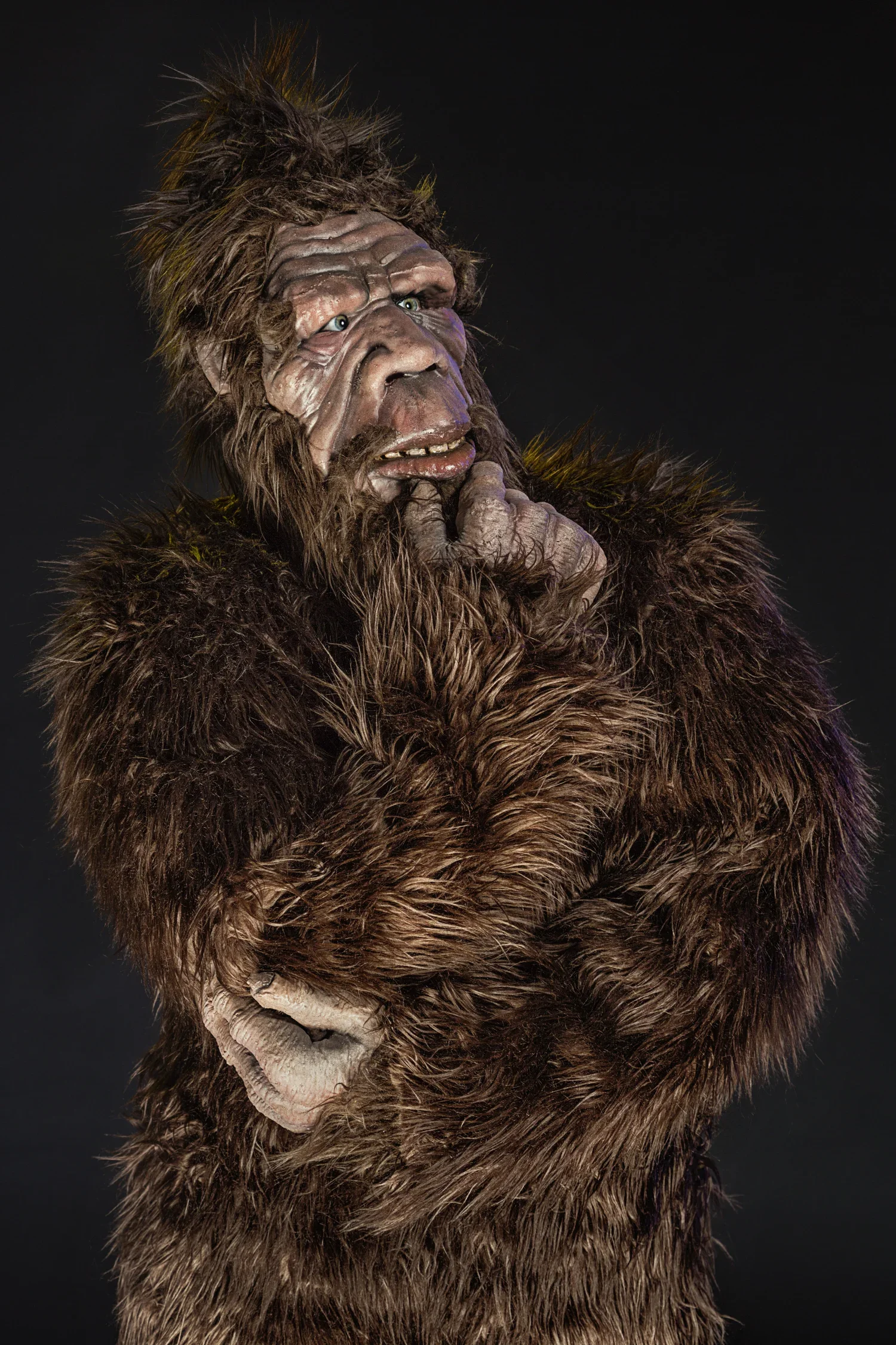A person dressed as a bigfoot or sasquatch creature with brown fur and detailed makeup, standing against a black background.