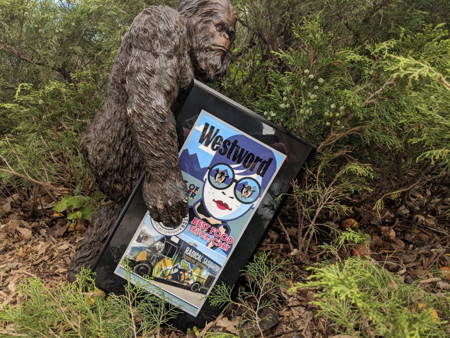 A gorilla sculpture holding a framed poster for Westword magazine, featuring a cartoon woman with sunglasses, surrounded by plants and shrubbery.