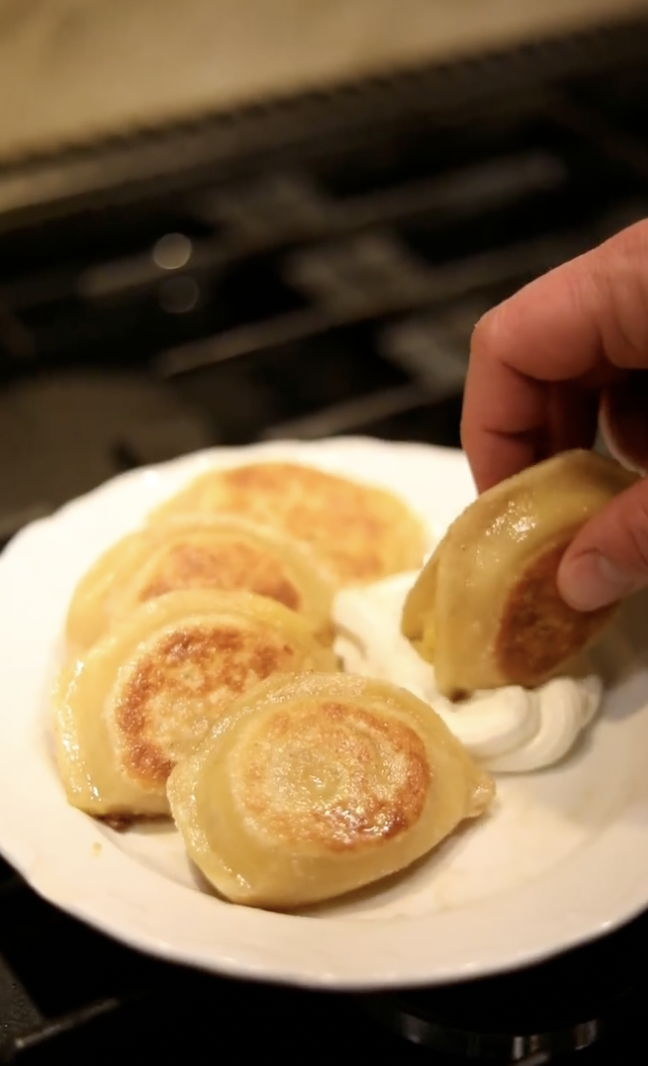 Close-up of a white plate holding five golden-brown pan-fried dumplings with a hand picking up one dumpling. The background is a dark stovetop.