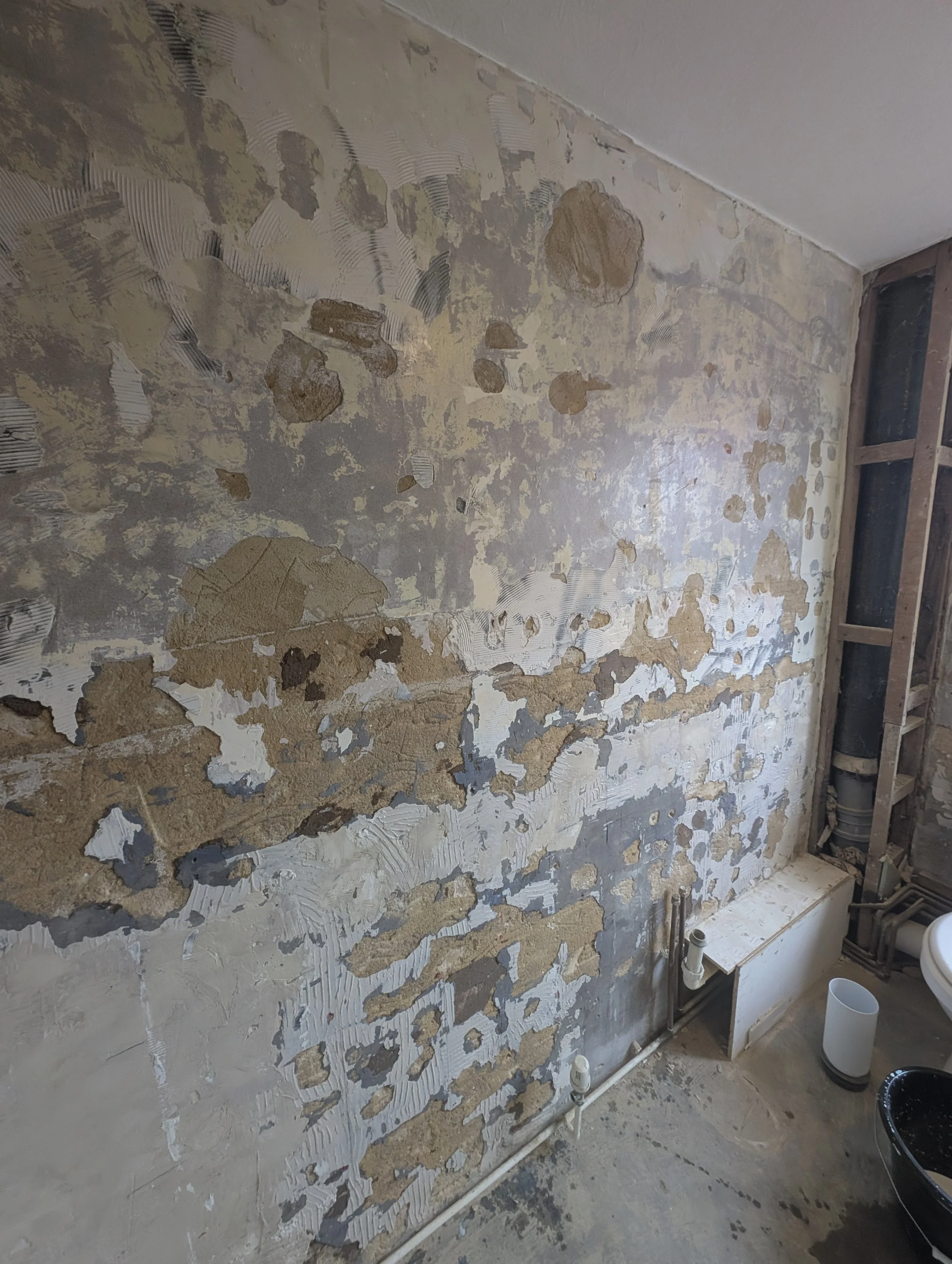 Interior wall under renovation with old plaster and drywall patches, exposed pipes, and construction materials on the floor.