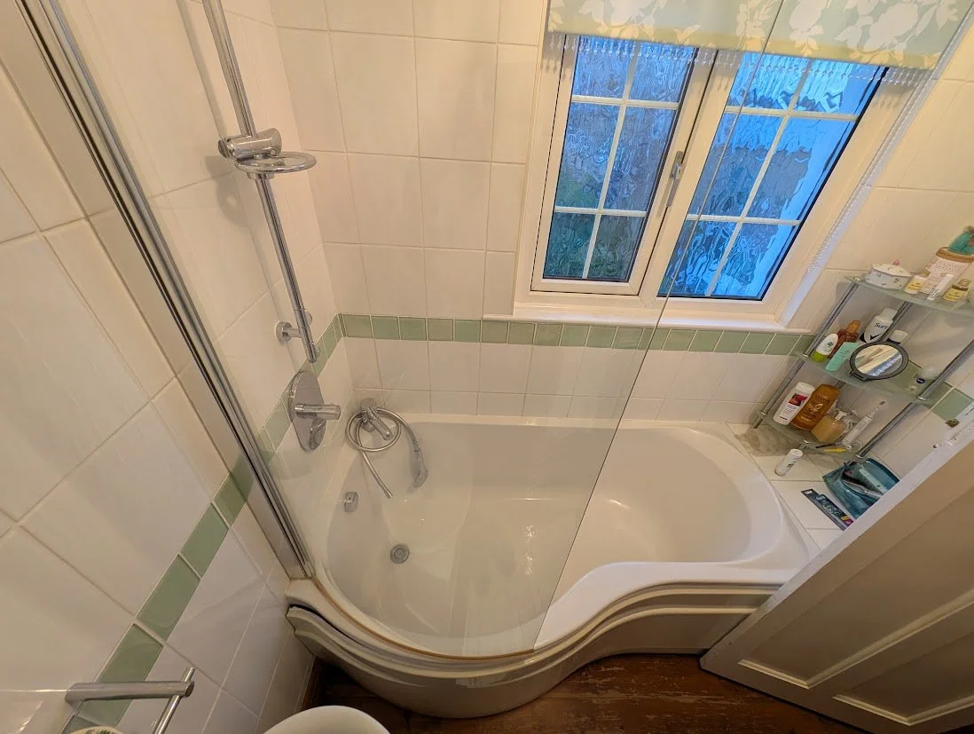 Small bathroom with a curved corner bathtub, a glass shower enclosure, and a window with frosted glass. There are shelves with toiletries beside the bathtub.