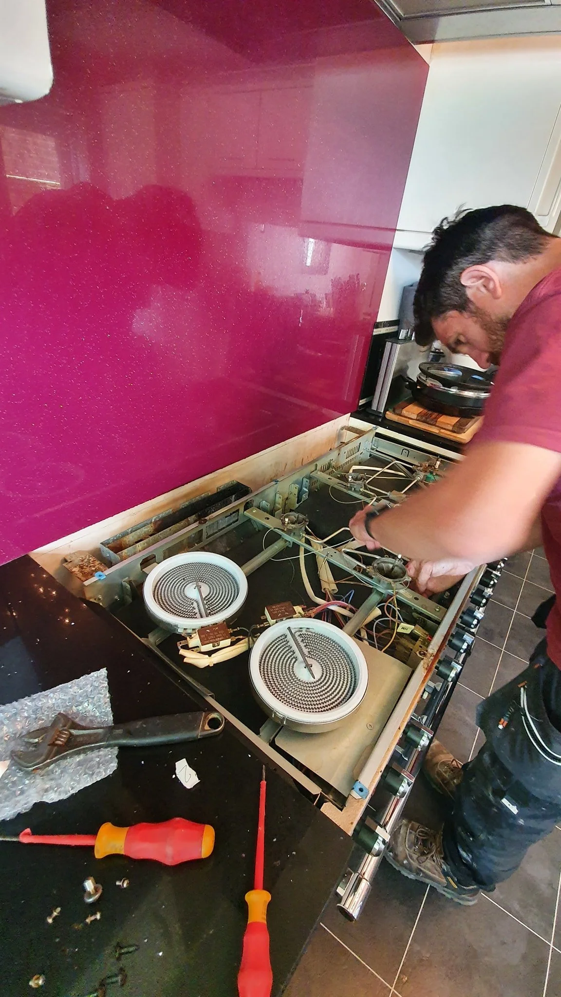 Man repairing or working on a broken electric stove, with the cooktop removed exposing the internal heating elements and wiring, on a black countertop in a kitchen.