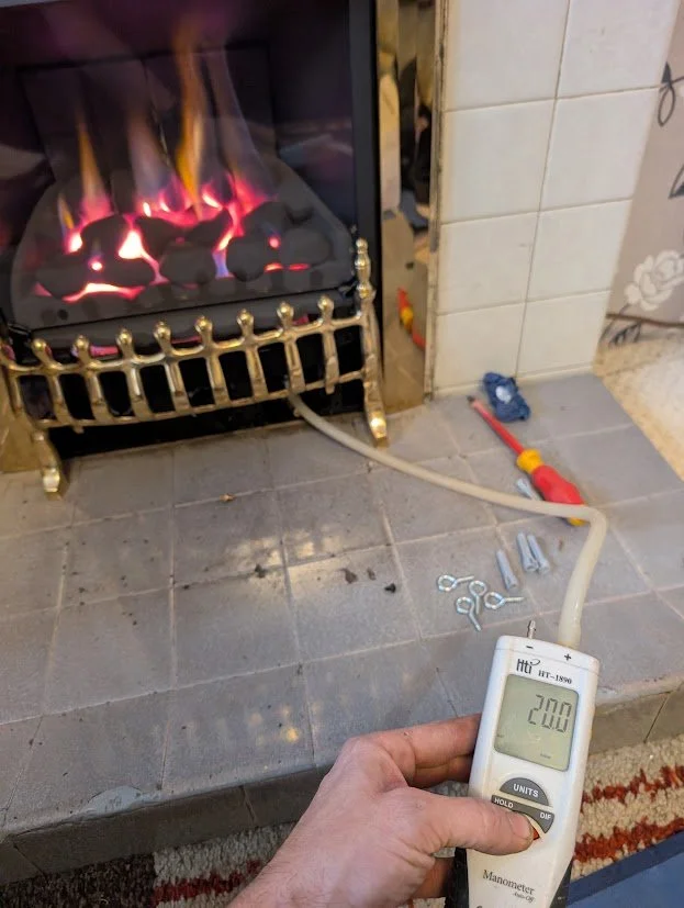 Fireplace with flames on gas logs, hand holding a manometer reading 200, with tools on the floor nearby.