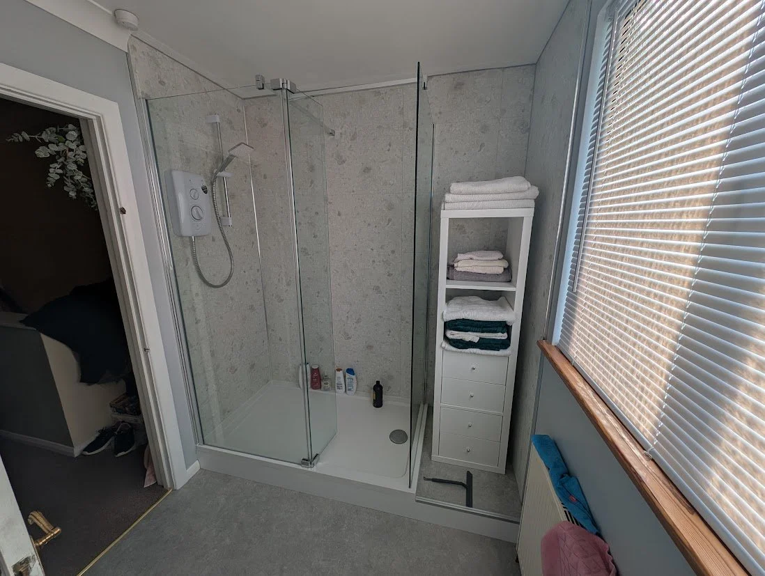 Bathroom with a glass shower enclosure, folded towels on a white shelf, and a window with blinds