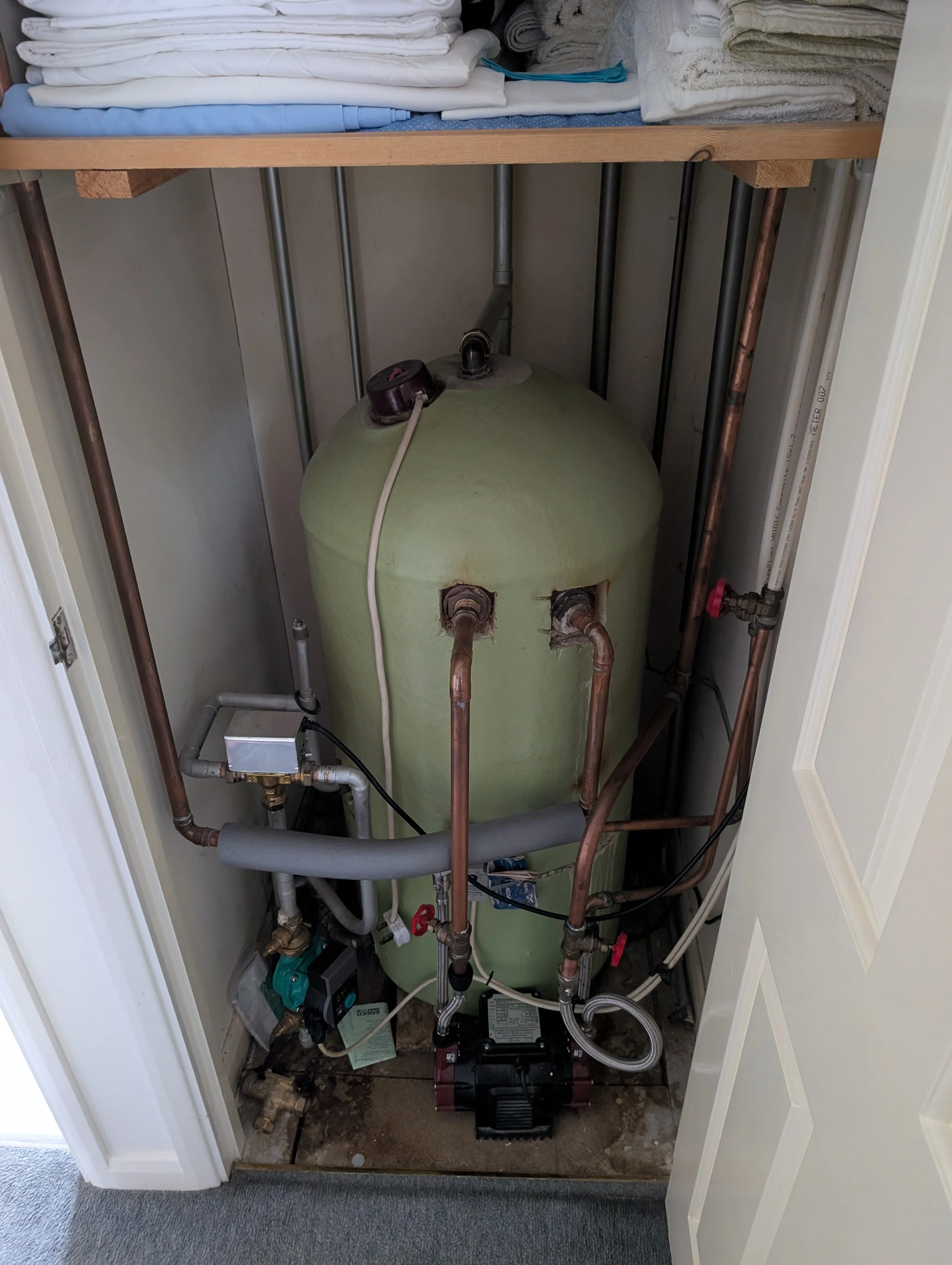 An open closet with an electric water heater, pipes, and a pump installed inside. The top shelf holds neatly folded towels and linens, while the bottom area contains plumbing components, pipes, and electrical wiring.