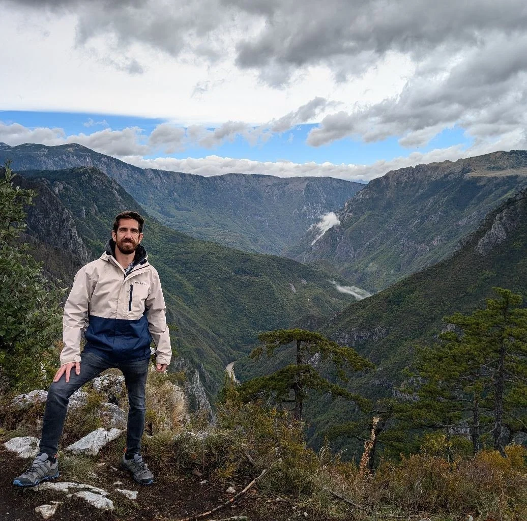 A man standing on a rocky ledge overlooking a lush green canyon with mountains in the background, cloudy sky overhead.