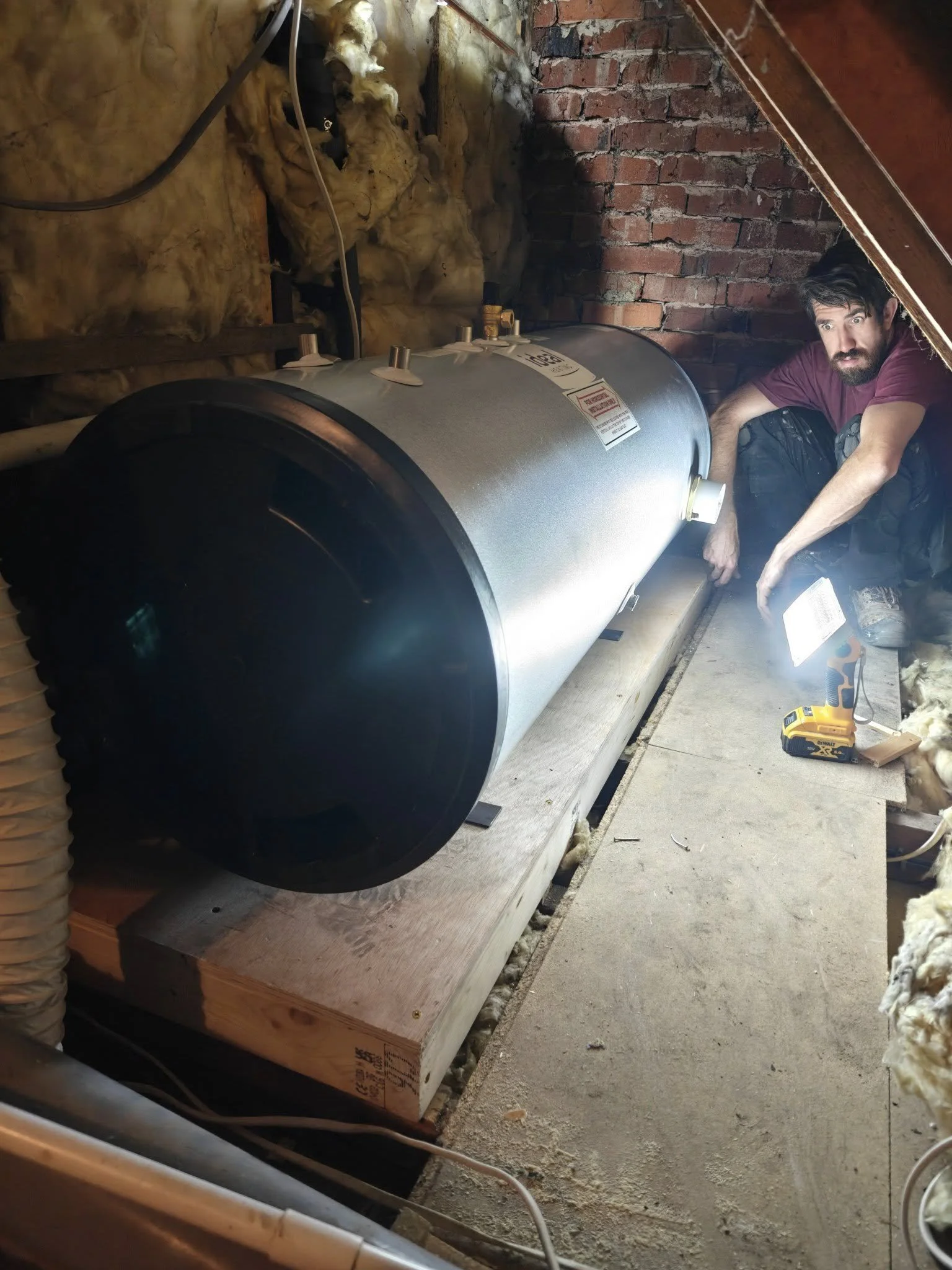 A man kneeling next to a large cylindrical tank in a crawl space with wooden flooring, insulation on the walls, and brick wall in the background, holding a tool and working on the tank.