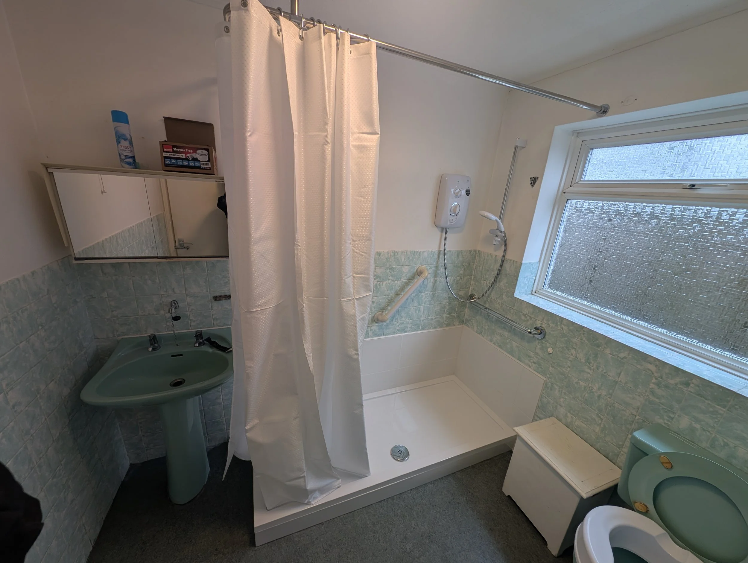 A small bathroom with a green sink, a shower with a white curtain, a window with frosted glass, and a green toilet. The walls are tiled halfway up with light blue and white tiles, and there is a mirror above the sink.