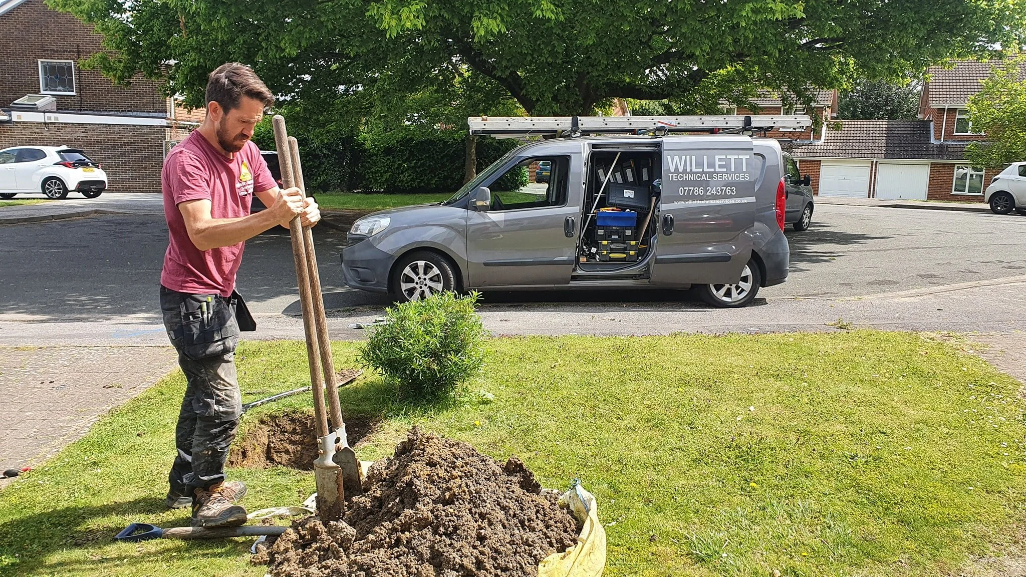 A man digging a mains water service pipe in a residential yard, with a garden tool in hand, near a small shrub and a pile of soil, with a service van parked on the street behind him.