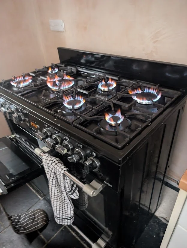 Black gas stove with six lit burners producing blue flames, with an oven door slightly open and a towel hanging from the handle.