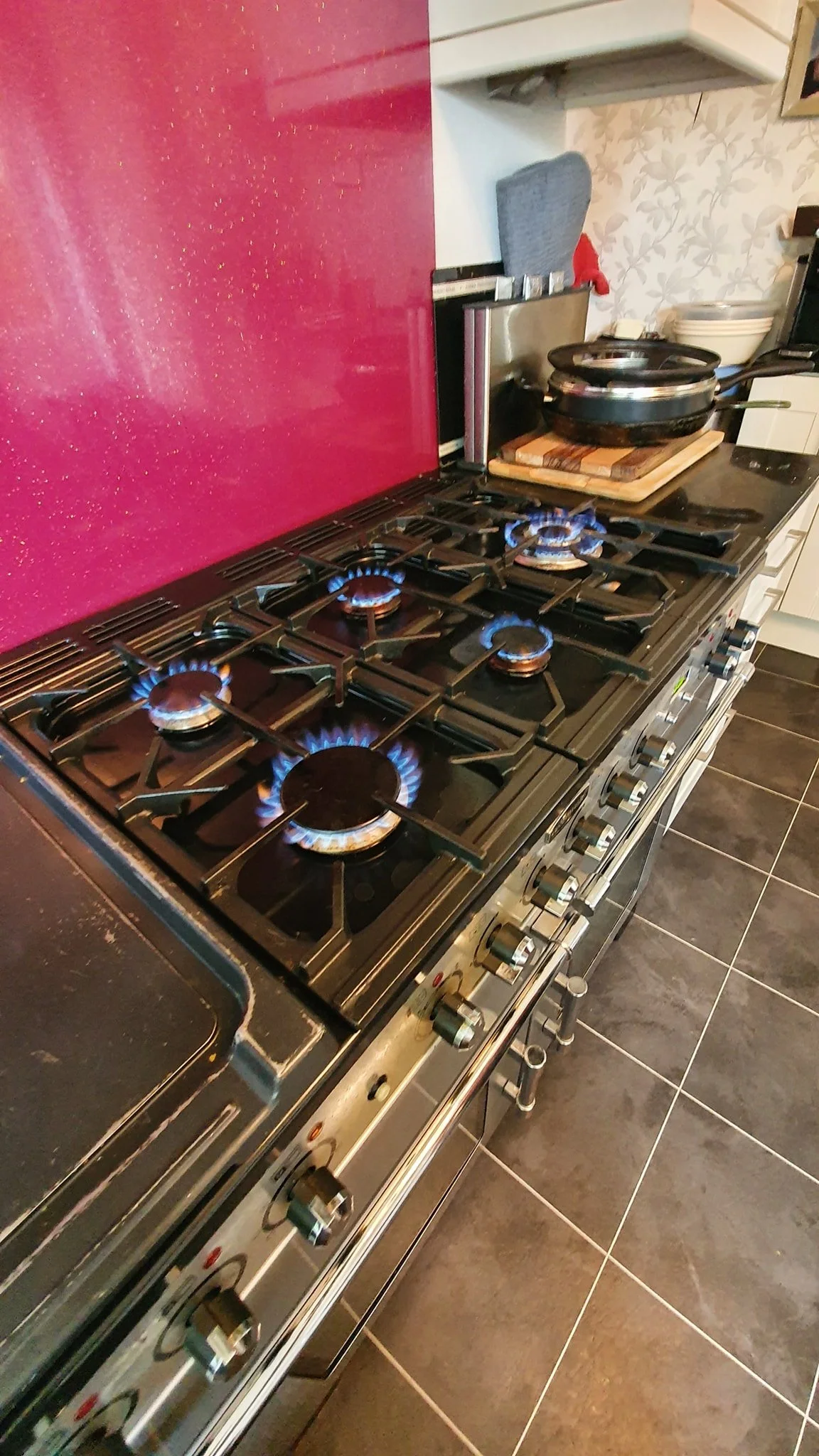 A stainless steel gas stove with five burners turned on and blue flames, situated in a kitchen with black tiled floor and a pink backsplash.