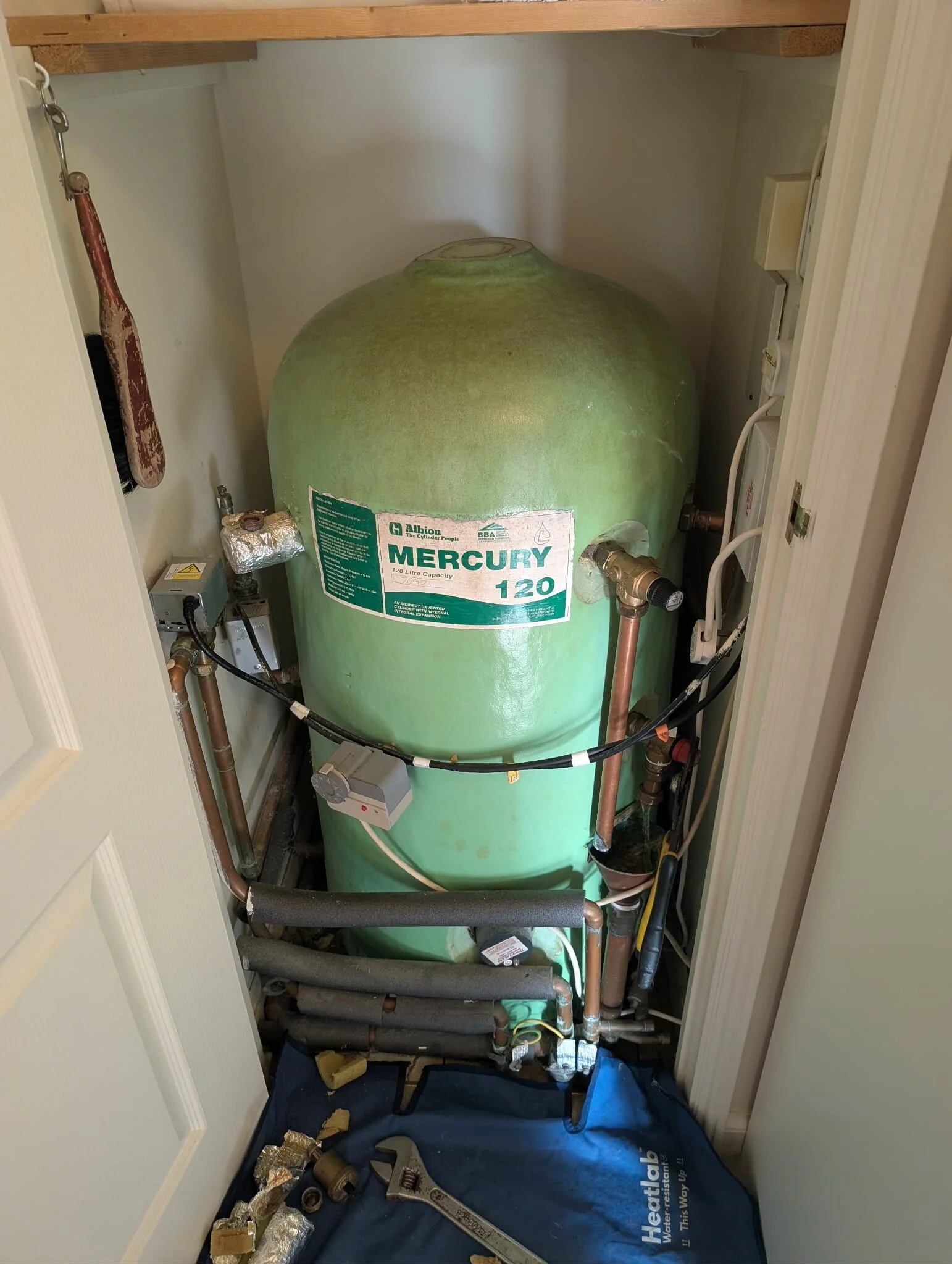A green Mercury water heater tank labeled 120 liters, installed in a small closet with copper pipes connected to it, with tools and insulation materials on the floor.