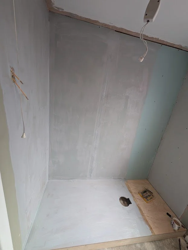 Room with unfinished drywall, electrical wires hanging from the ceiling, a hole in the floor, and a piece of plywood with tools and a cup on it.