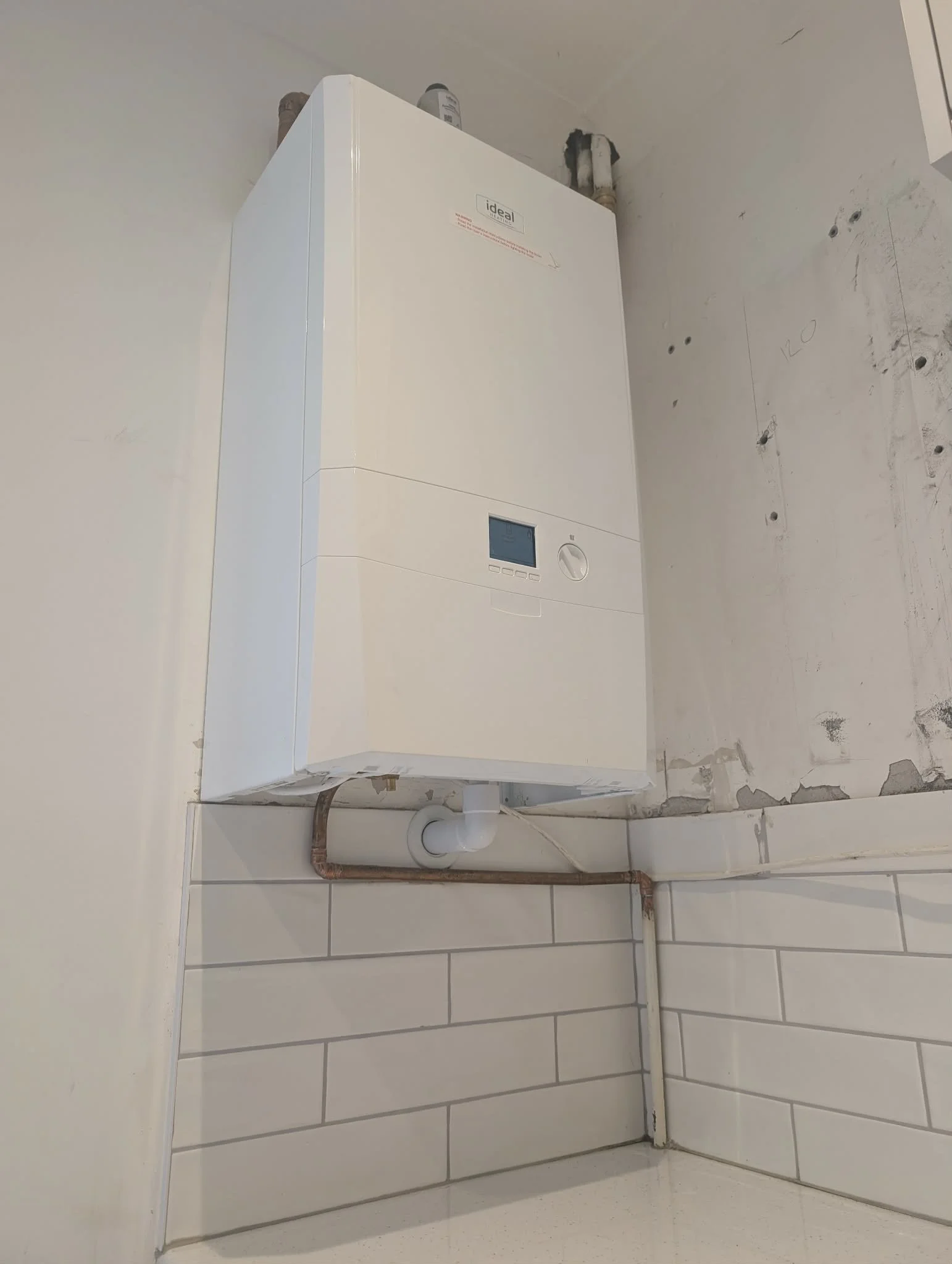 A white tankless water heater mounted on a wall in a laundry room, with exposed pipes and unfinished wall sections visible.