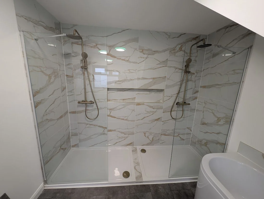 Double shower with beige and white marble tiles, two overhead rain shower heads, and a partial view of a curved white basin in a modern bathroom.