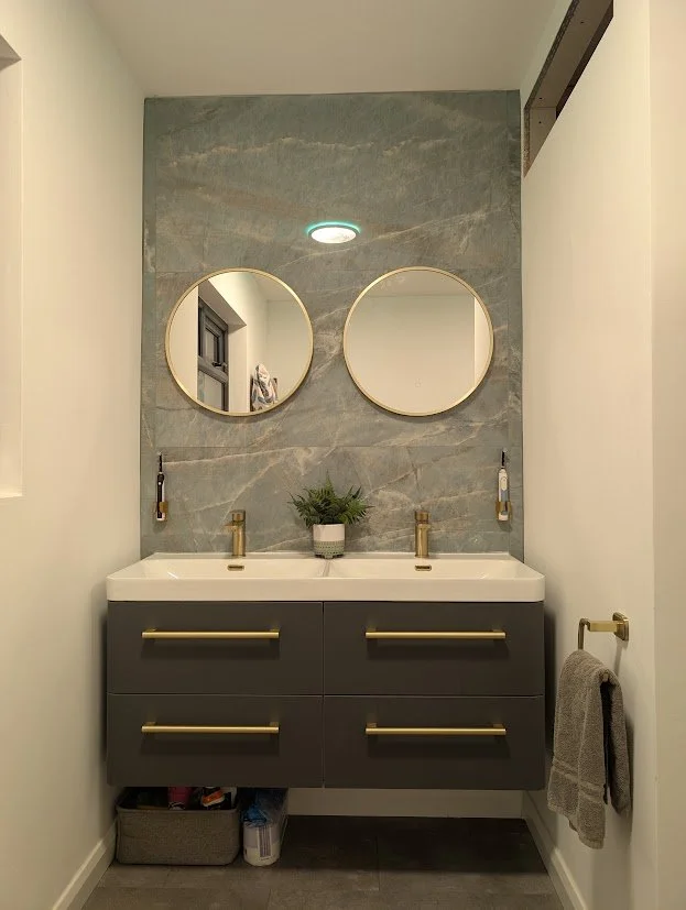 A modern bathroom vanity with a double sink, gray drawers with gold handles, two round mirrors on a marble backsplash, a potted plant, and a wall-mounted towel ring with a gray towel.