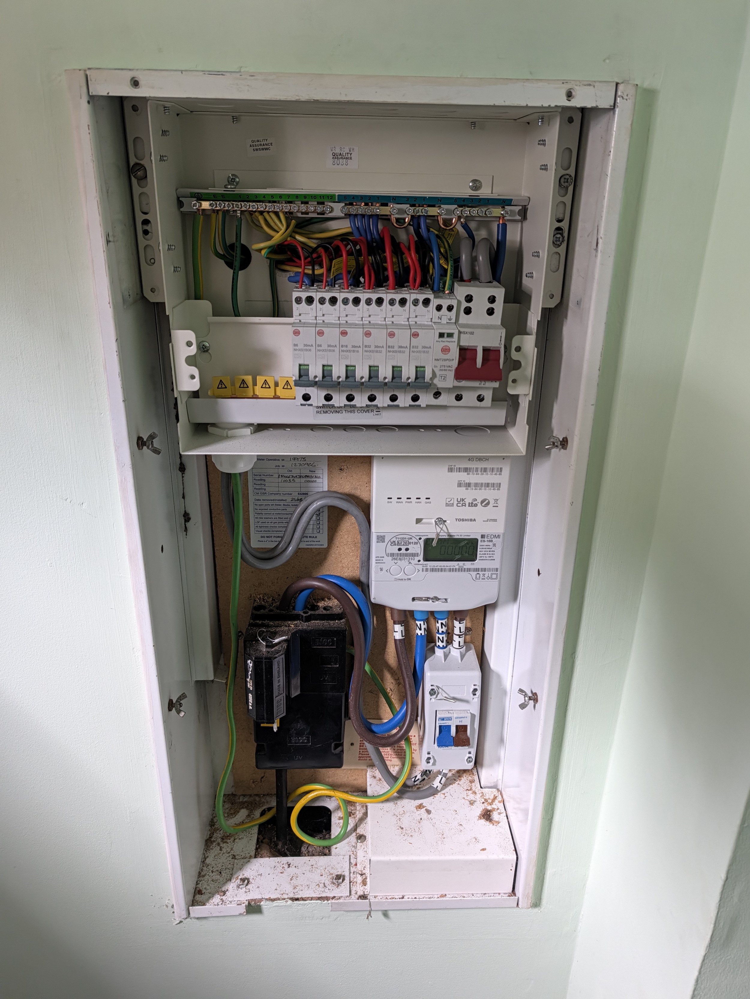 An electrical breaker panel with various colored wires connected to circuit breakers inside a white metal box installed in a wall.