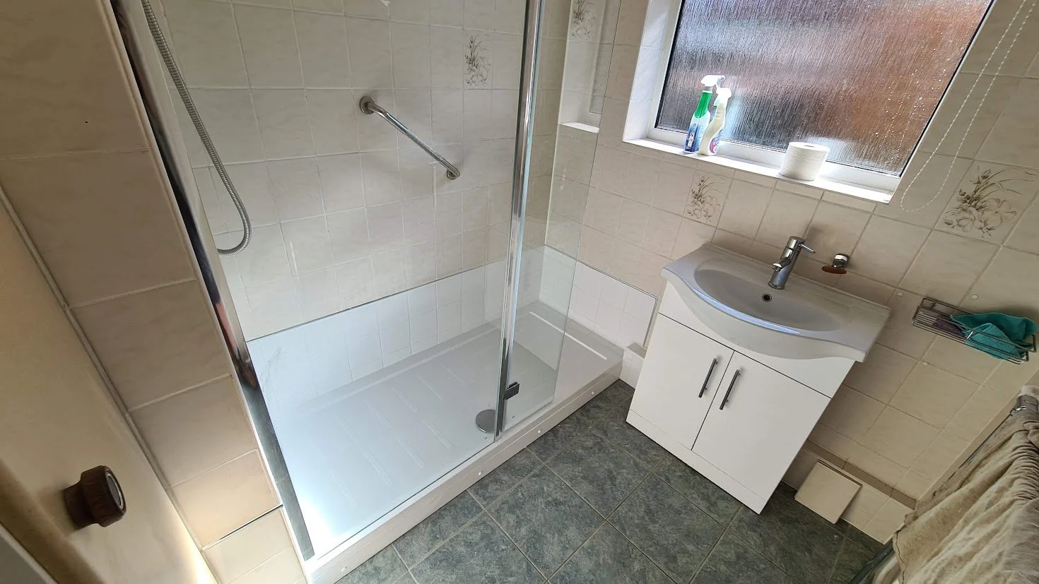 Small bathroom with a shower enclosure, a white sink cabinet, and a frosted window with cleaning supplies and a roll of toilet paper on the windowsill.
