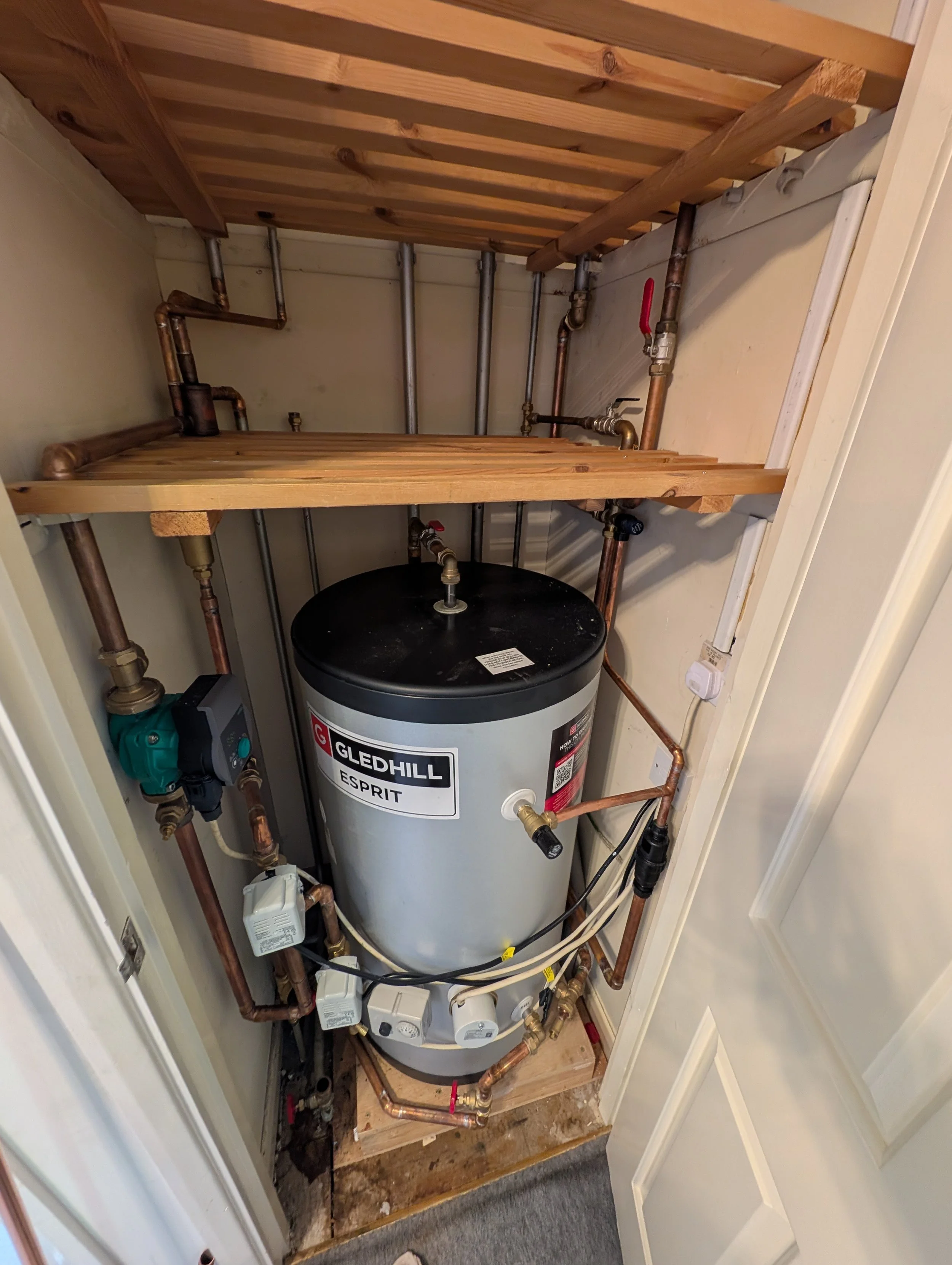 A utility closet with an exposed water heater, copper piping, and some electrical components, with a wooden platform above the water heater.