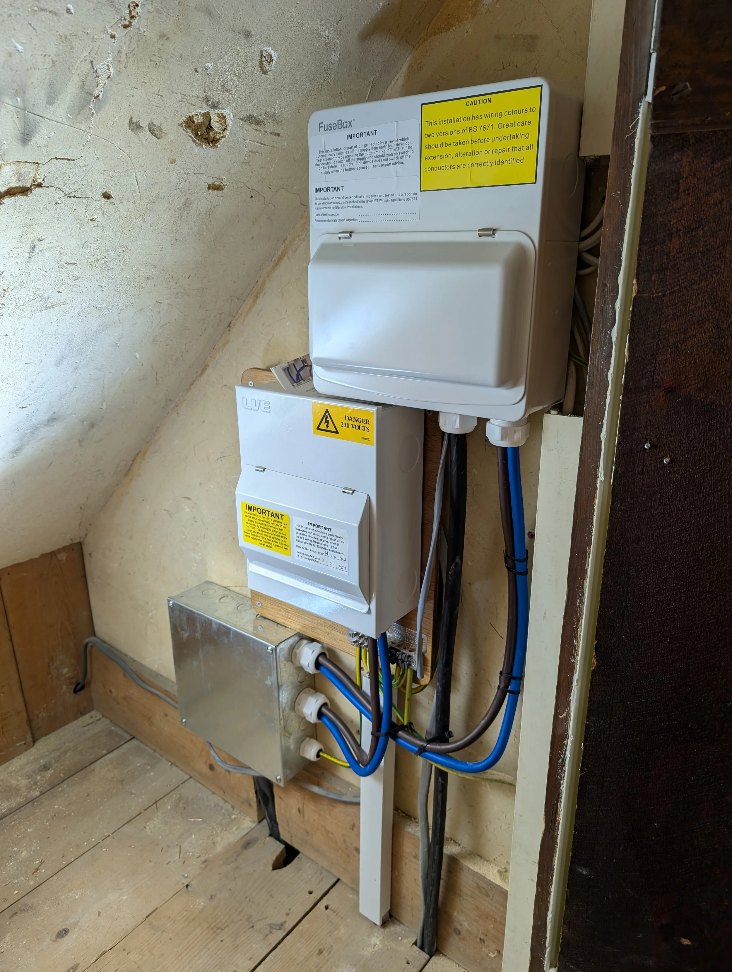 Electrical fuse and circuit box mounted on a wall in a rustic interior, with wiring and cables connecting to the units. The fuse box has a caution sign warning of 230 volts and an important label with safety information.