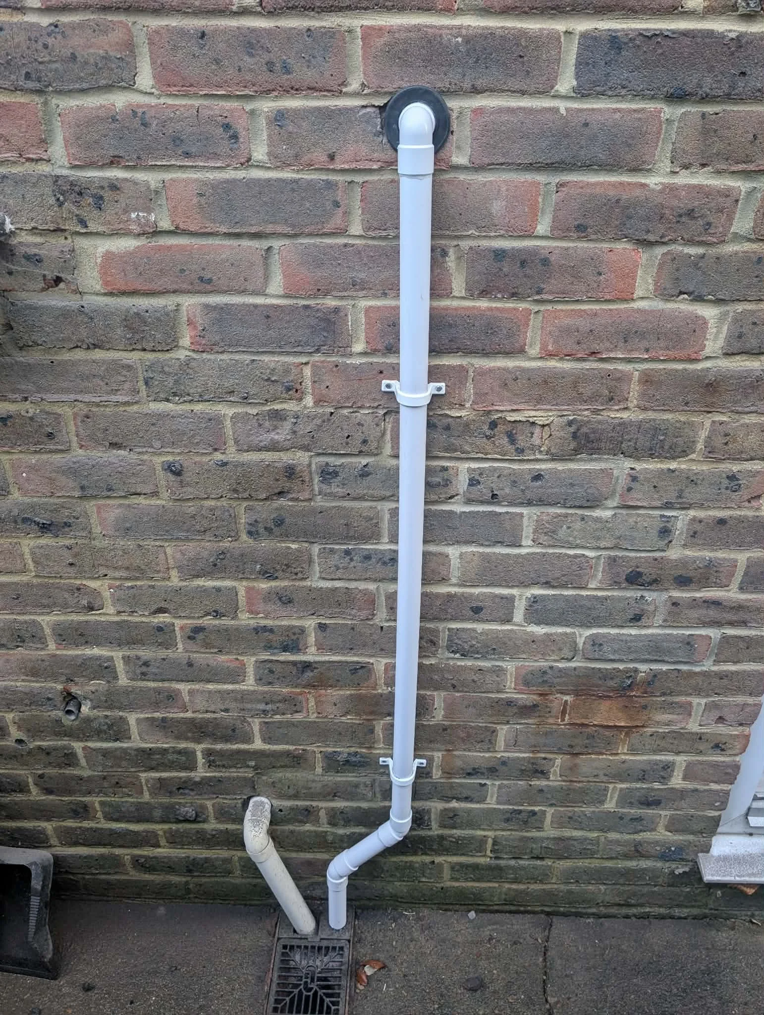 White PVC pipe drainage system mounted on a brick wall, extending from ground to near the top of the wall with an elbow pipe at the bottom connecting to a drain.