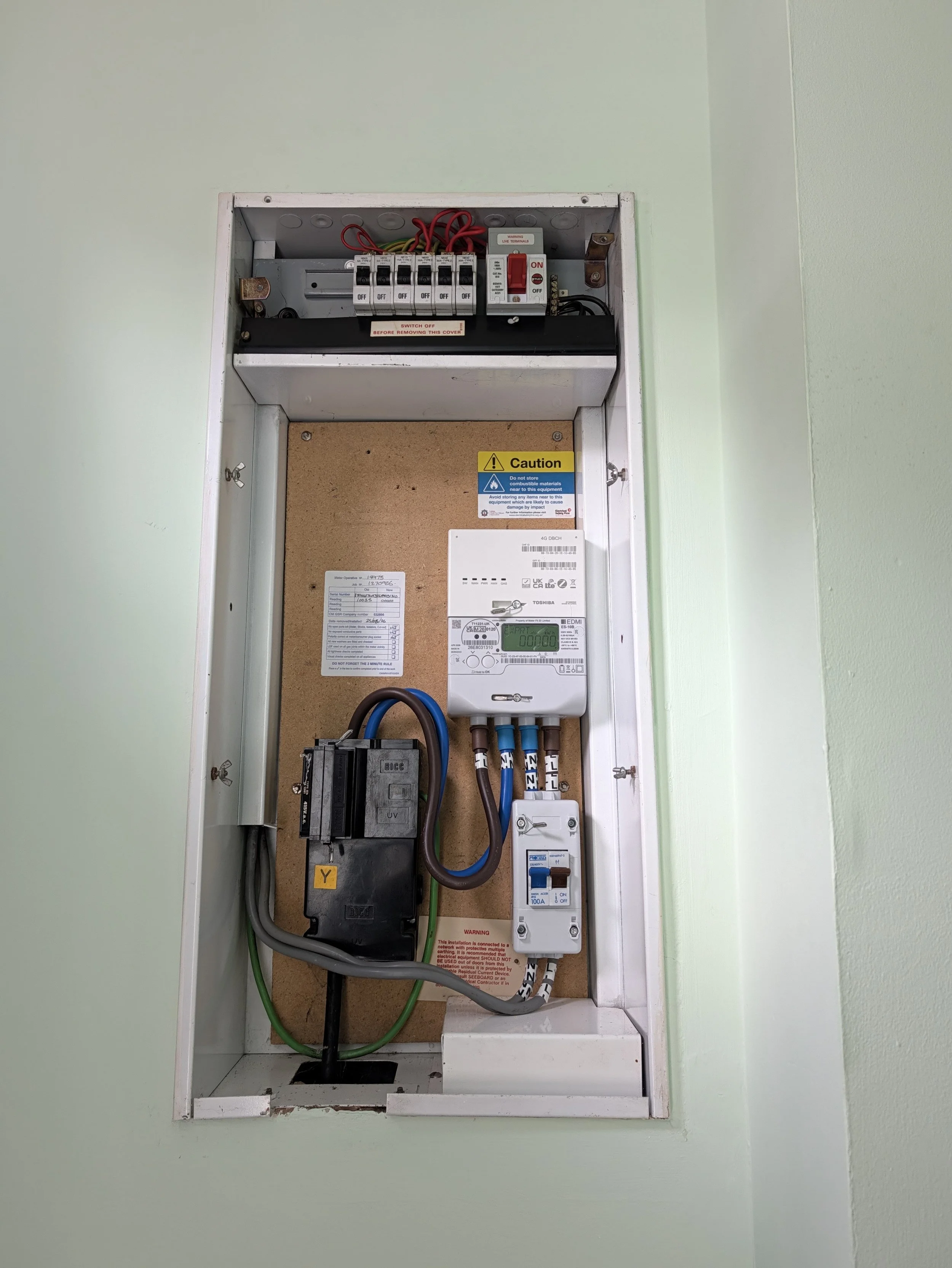 Electricity meter and circuit breaker inside an electrical panel on a green wall.