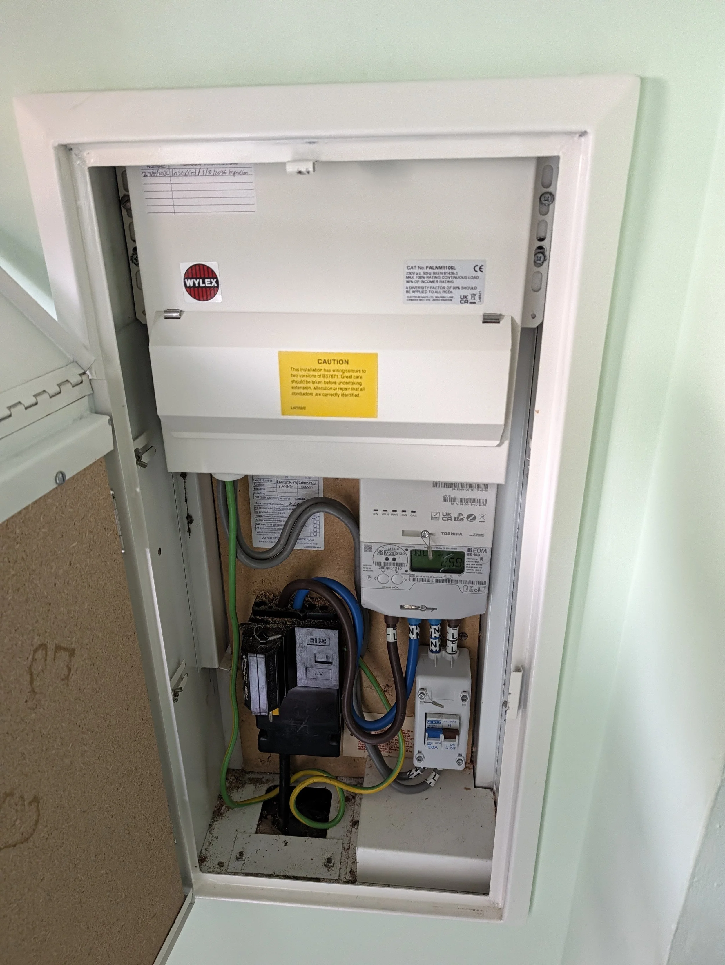 Electrical panel with various components, wires, and labels inside a wall-mounted box, with some green and yellow wiring connected.