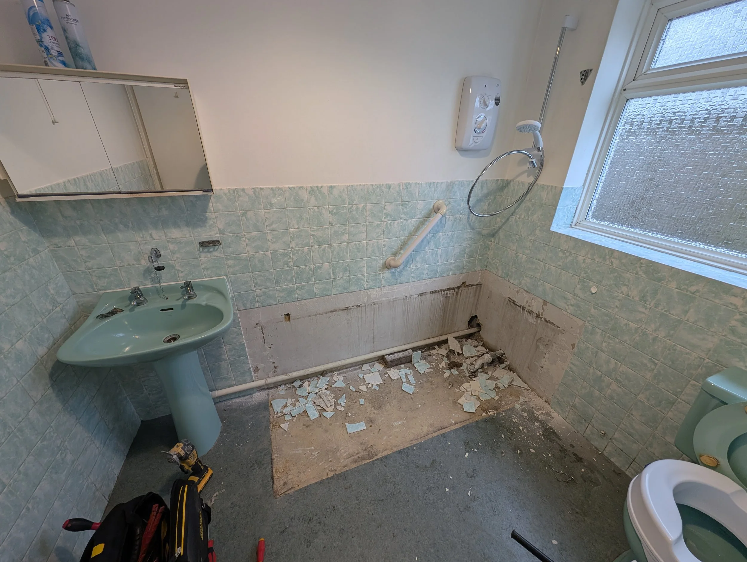Bathroom in renovation with exposed wall and floor sections, dusty debris, a sink, mirror cabinet, showerhead, and a window.