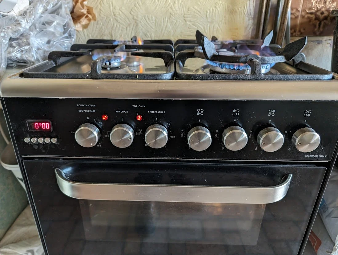 A black and stainless steel gas stove with six burners, two of which are lit, and an oven below. The control knobs are on the front panel.