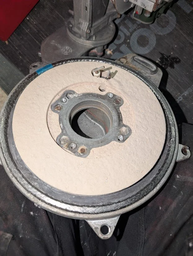 A metal mechanical part mounted on a circular base, with a central hole and six bolt holes around the center, covered with beige insulation or padding, placed on a dark work surface.
