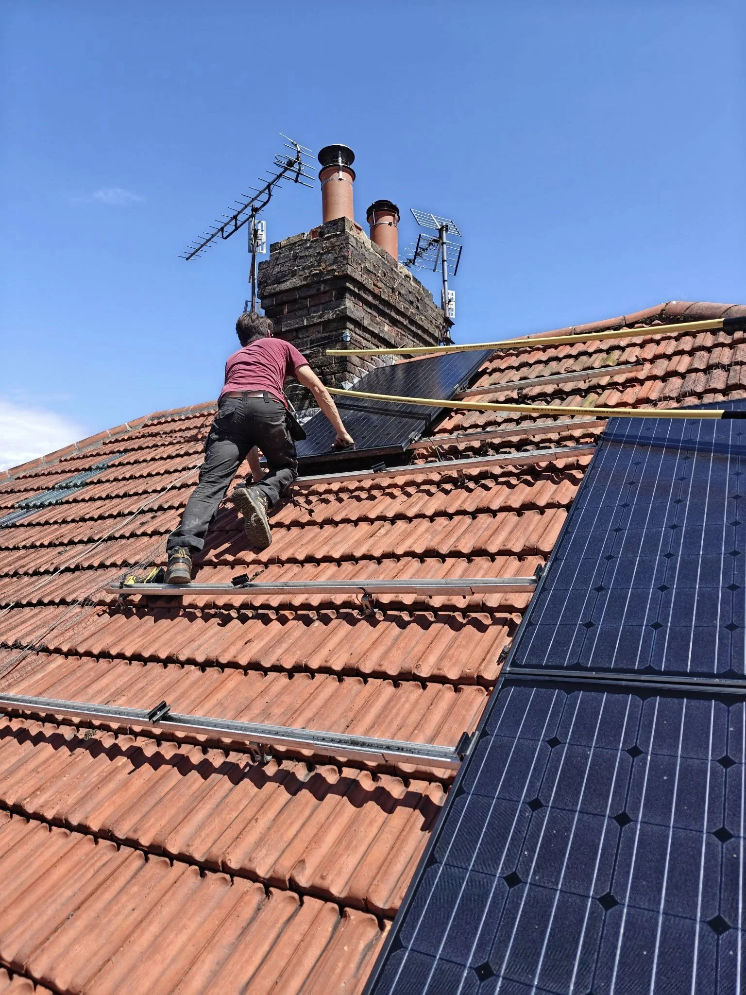 A worker installing solar panels on a sloped rooftop with red tiles, with a blue sky in the background. The worker is climbing on the roof and holding a solar panel while attaching it to mounting rails. There are some antennas and chimney stacks on the roof.