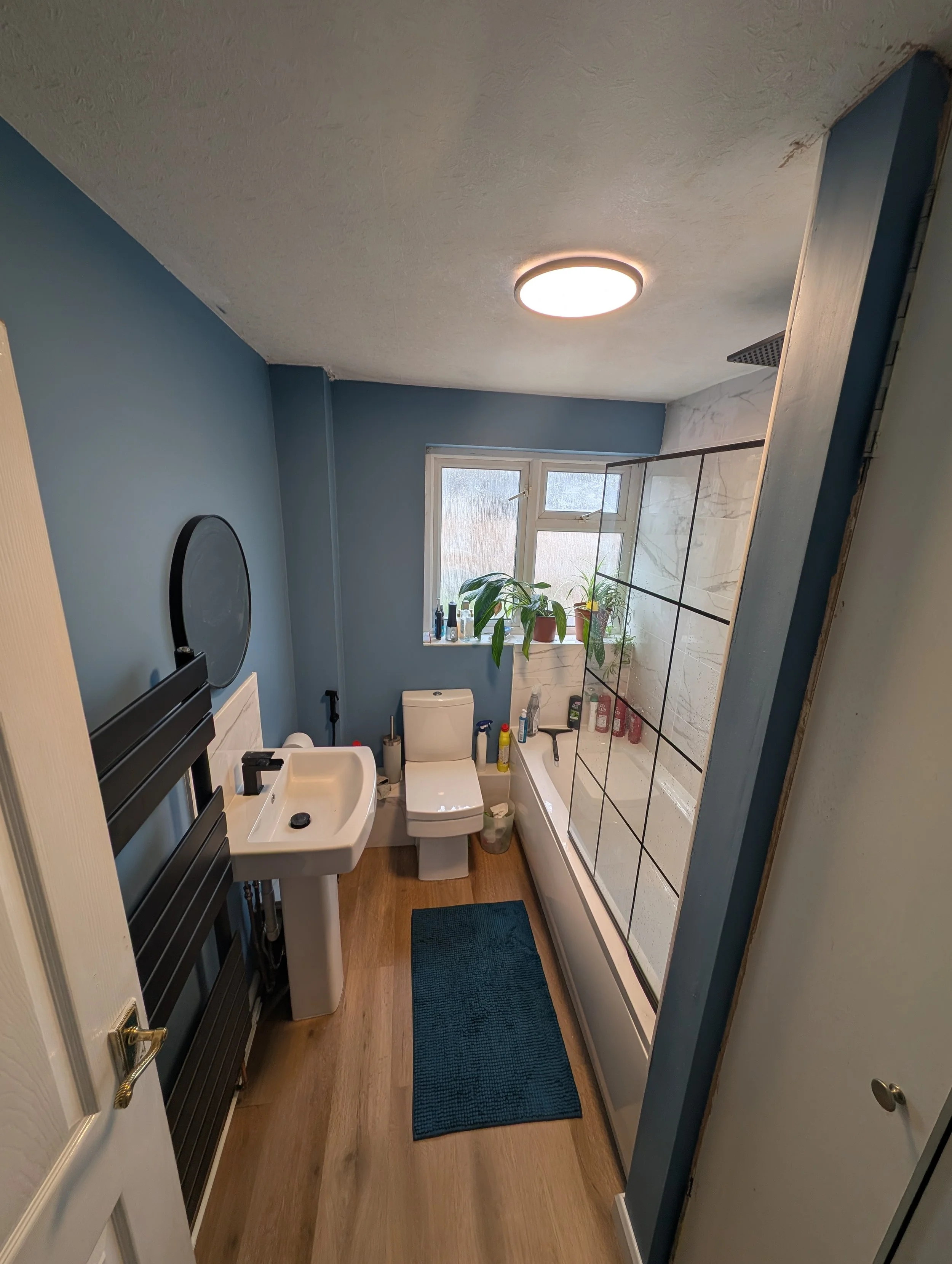 A bathroom with blue walls, a white toilet, a small white sink, a shower with glass doors, and a window with plants on the sill. There are various toiletries on the window ledge and shower wall, a blue bath mat on wooden flooring, and a radiator on the left.