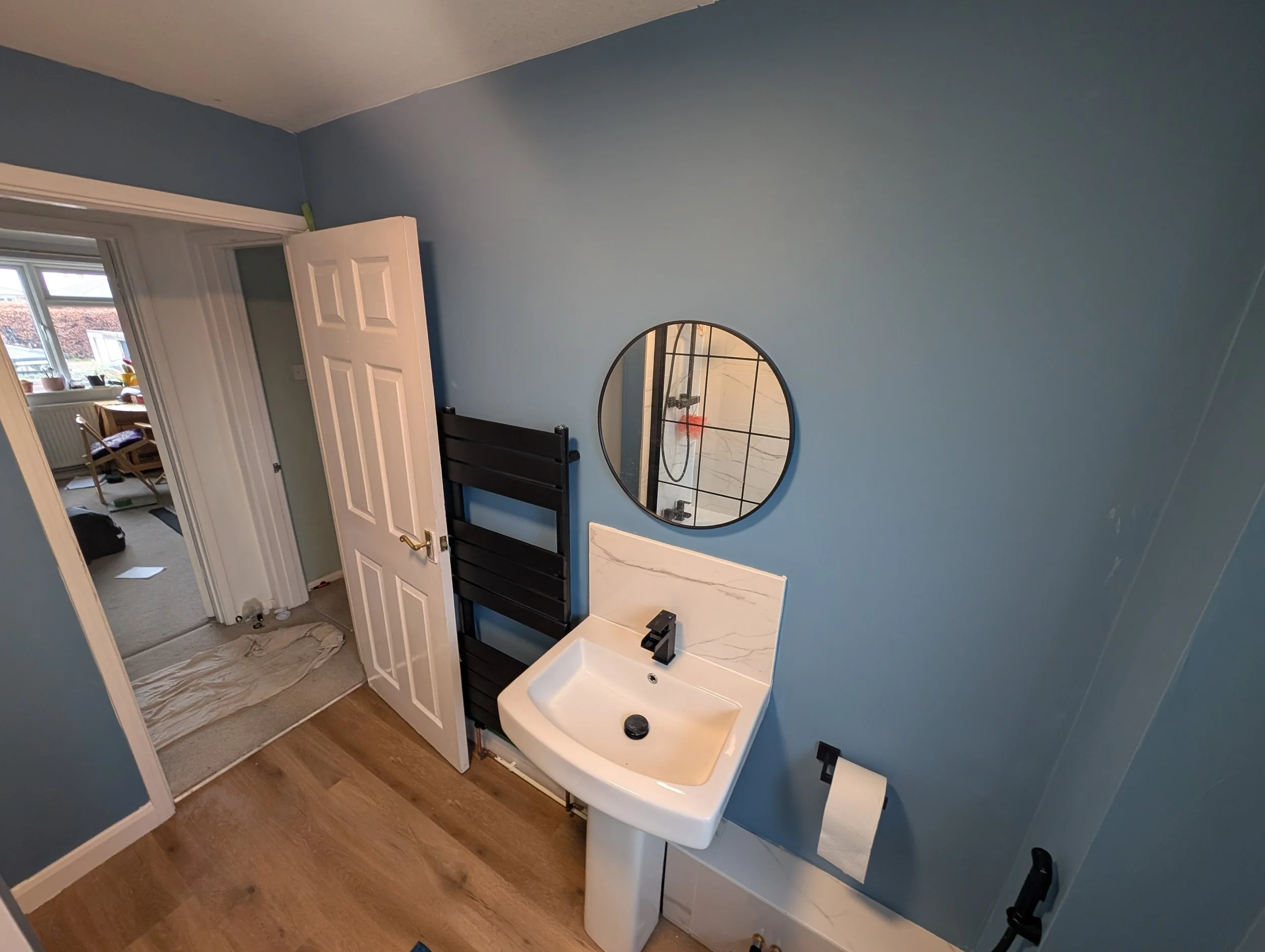 A small bathroom with blue walls, a wall-mounted round mirror, a white sink with black faucet, and a black towel rack. An open door leads to another room with desk, chairs, and large window.