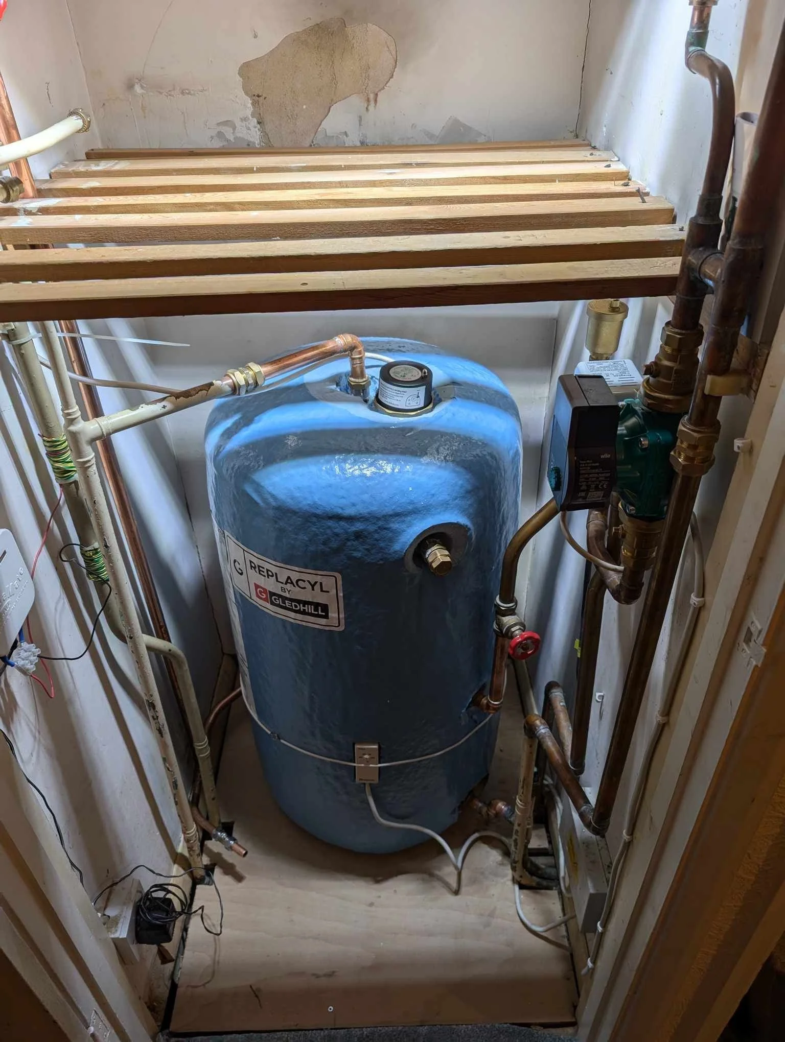 A water heater and plumbing in a small utility closet with unfinished walls and a wooden shelf above.