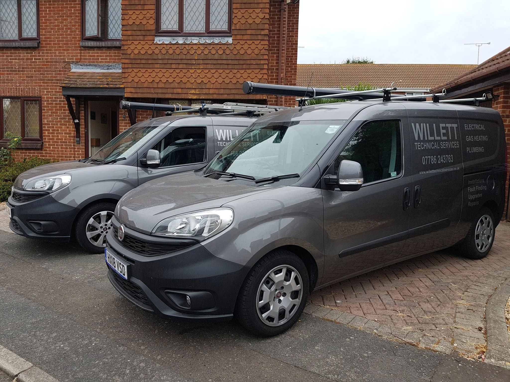 Two gray company vans parked in front of a brick building, with the company name 'WILLET' and services listed on the side, including electrical, gas heating, and plumbing.