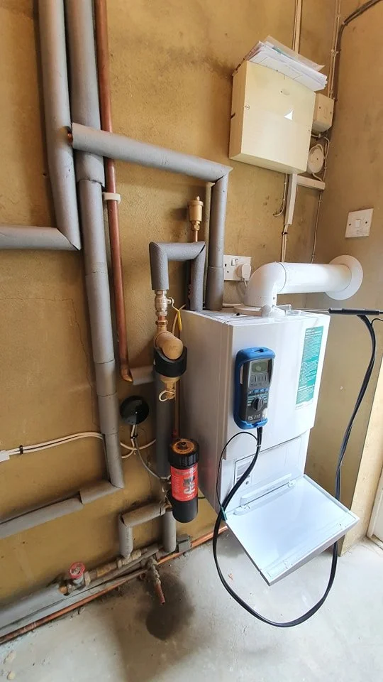 A utility or boiler room with pipes, a water meter, and a boiler unit connected to a vent pipe, with wires and a control device attached.