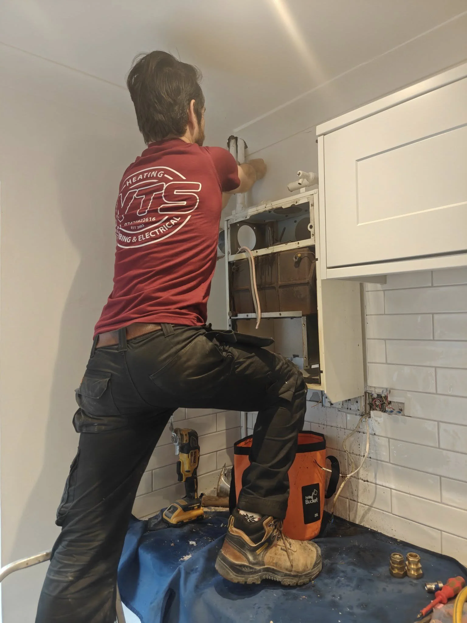 A handyman in work boots installing or repairing an over-the-range microwave or cabinet in a kitchen, with tools and a bucket nearby.