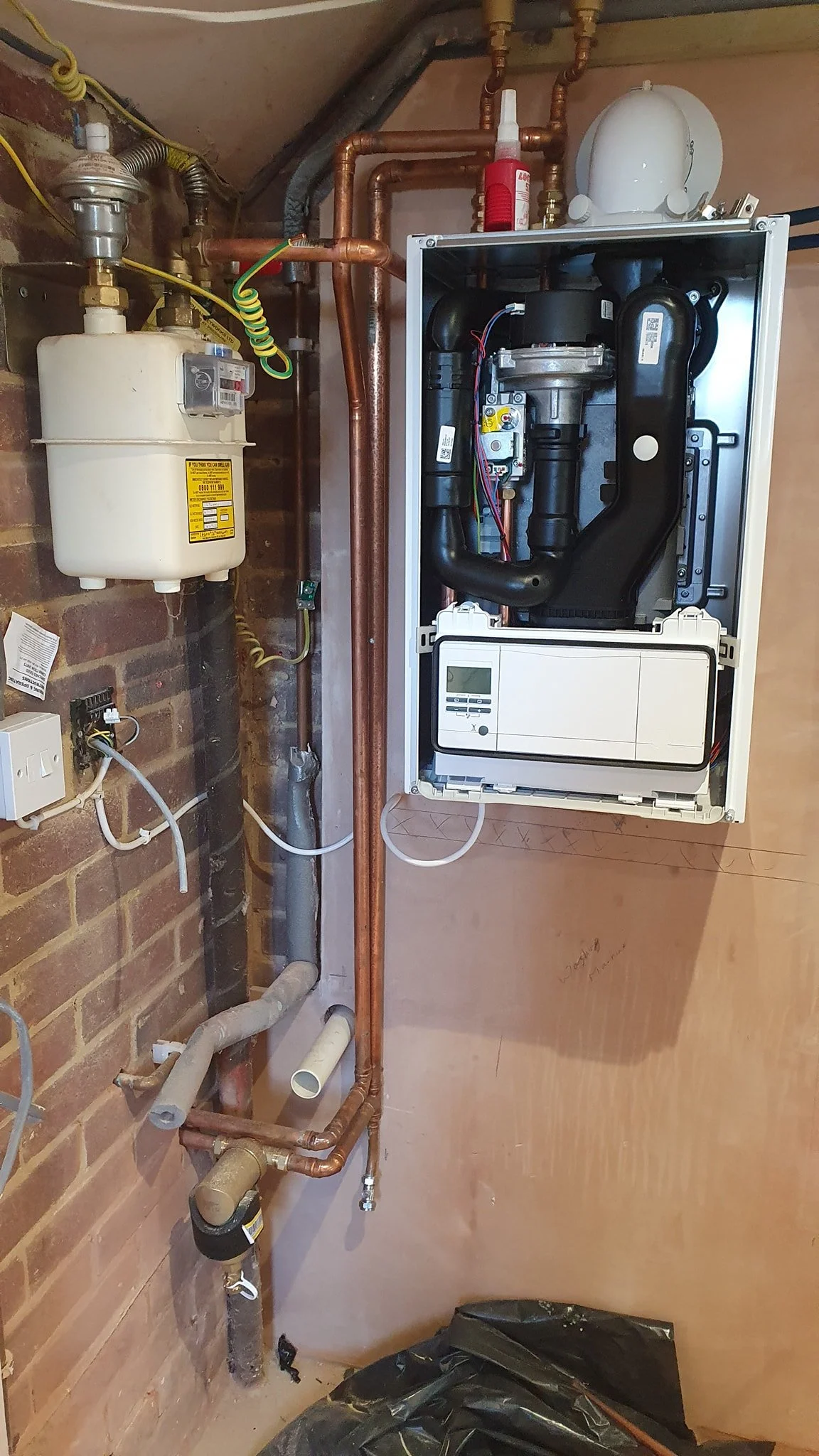Installation of a new boiler system with copper and black pipes, electrical components, and a control panel, mounted on a wood-paneled wall in a utility room.