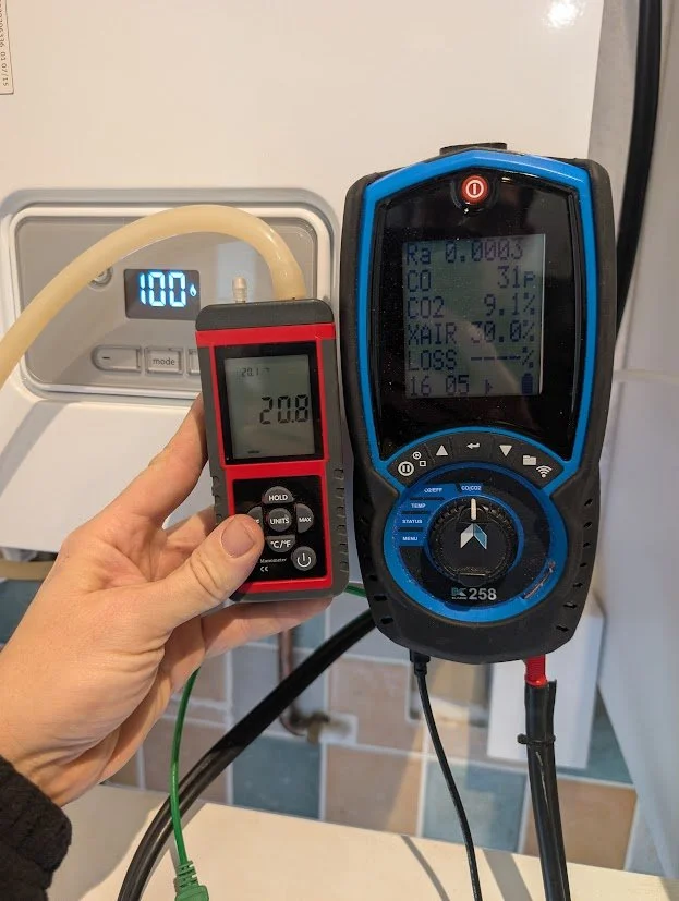 A person's hand holding a digital temperature and humidity meter showing 20.8°C, in front of a climate control device displaying 100°F temperature.