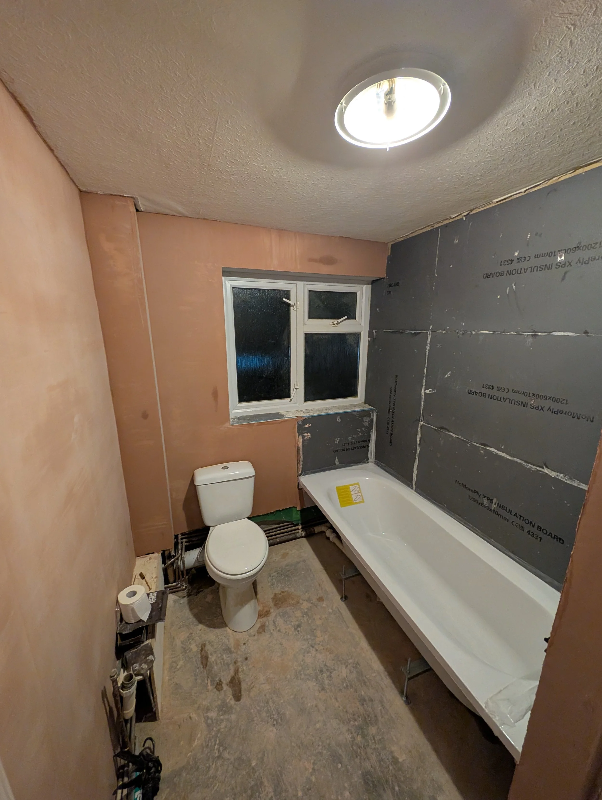 A bathroom under renovation with a toilet, unfinished wall with insulation, a window, and a bathtub with protective plastic, a ceiling light fixture, and exposed pipes on the floor.