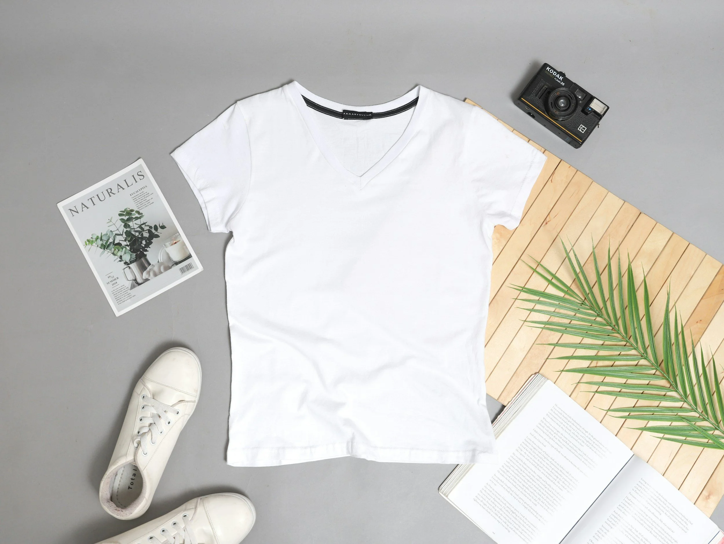 Flat lay of a white t-shirt, white sneakers, a magazine, an open book, a palm leaf on a wooden board, a black film camera, and a magazine titled 'NATURALIS' on a gray background.